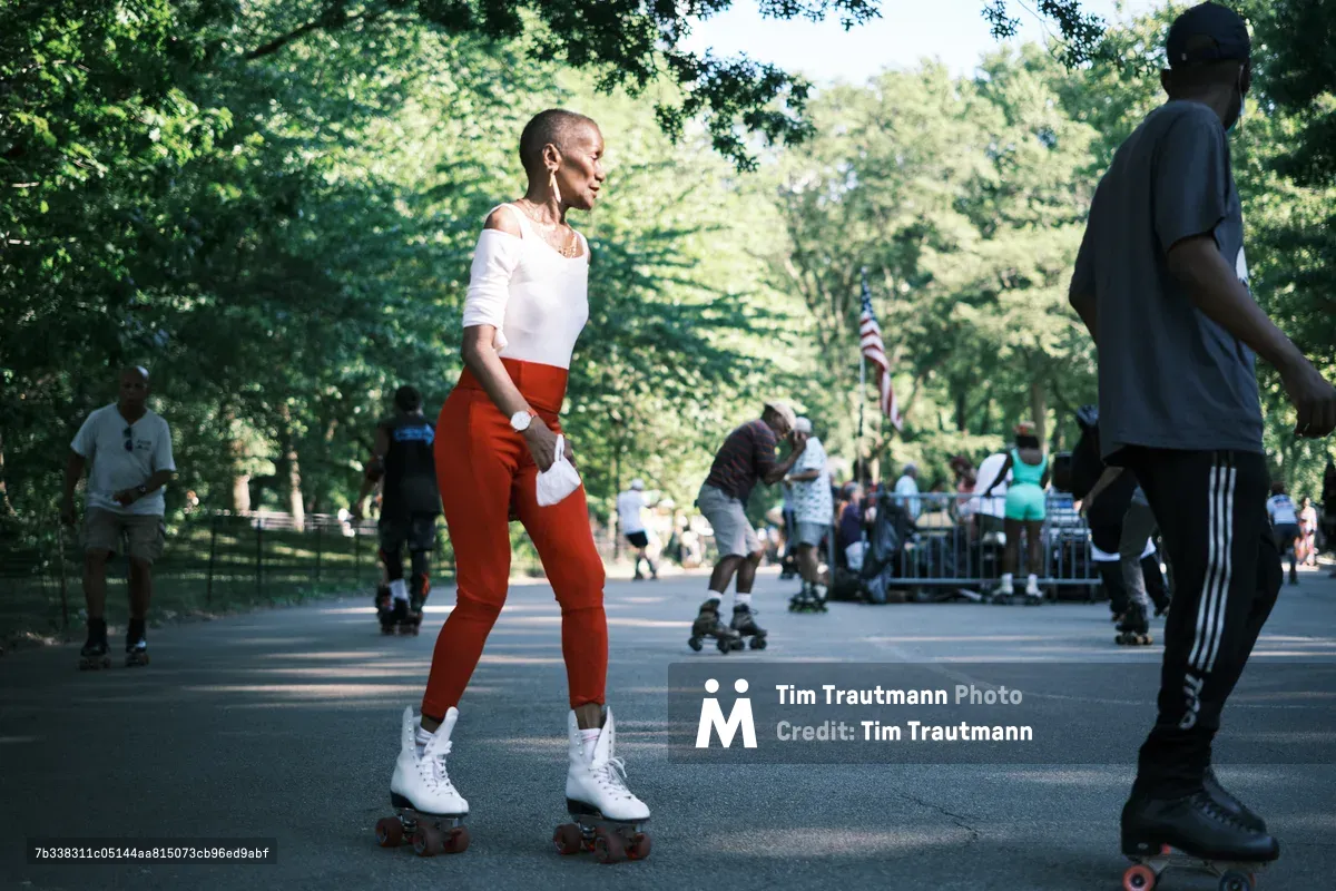 A woman in striking red pants and white off-shoulder top glides gracefully on white roller skates along Central Park's tree-lined pathways, her confident posture and gentle smile embodying the joy of summer recreation. The dappled sunlight filters through the lush green canopy above, creating a nostalgic atmosphere as fellow skaters and park-goers blur softly in the background. Her athletic stance and focused expression capture a moment of pure freedom and movement, while the classic roller derby aesthetic brings vintage charm to this contemporary urban scene.