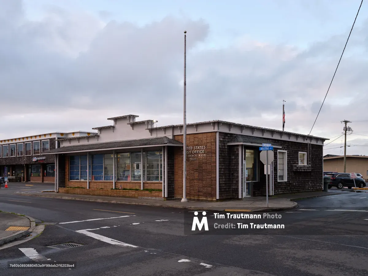 The Long Beach, Washington post office stands as a monument to mid-century civic architecture, its weathered cedar shingles and red brick facade anchoring a corner lot under brooding Pacific coastal clouds. The building's clean modernist lines are softened by the overcast light that blankets the small beach town, while wet asphalt reflects the muted tones of the sky above. A solitary flagpole pierces the gray expanse, and the quiet intersection speaks to the unhurried rhythm of this peninsula community where the Columbia River meets the sea.