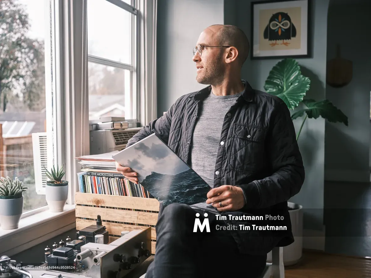 A bearded man wearing glasses and a quilted black jacket sits by a large window, examining vinyl records from a wooden crate. The cozy Portland home interior features plants, framed artwork, and natural lighting.