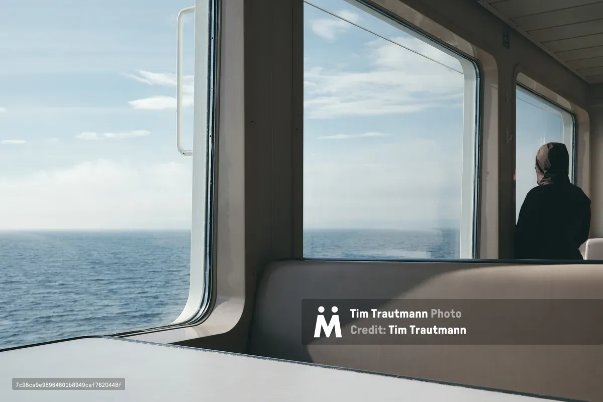 The interior of a Washington State Ferry passenger cabin, with large windows looking out over the calm blue waters of Puget Sound under a partly cloudy sky. A lone passenger wearing a dark jacket and head covering sits with their back to the camera, gazing out at the open water. Empty beige bench seating runs along the window row in the foreground.