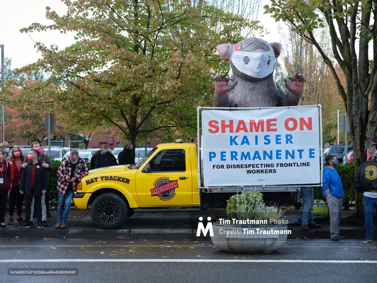 A towering inflatable rat wearing a surgical mask looms over a labor demonstration in Portland's Lloyd District, its claws raised menacingly beside a bright yellow truck bearing the message 'Shame on Kaiser Permanente.' Masked healthcare workers and union supporters cluster around the symbolic creature beneath autumn trees, their presence creating a powerful tableau of organized dissent. The overcast Oregon sky provides a muted backdrop to the bold red and blue protest signage, while fallen leaves scattered across the pavement hint at the season of change both literally and figuratively.