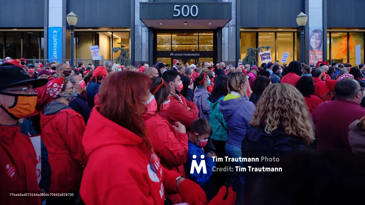 A dense crowd of healthcare workers, predominantly wearing red Kaiser Permanente scrubs and winter jackets, gathers in solidarity outside the Kaiser Permanente Tower at 500 Northeast Multnomah Street in Portland's Lloyd District. The sea of crimson creates a powerful visual statement against the modern glass and steel facade of the medical facility, while protest signs punctuate the crowd and evening light casts a determined atmosphere over the assembled nurses. Face masks worn by many participants underscore the contemporary healthcare context of their labor action.