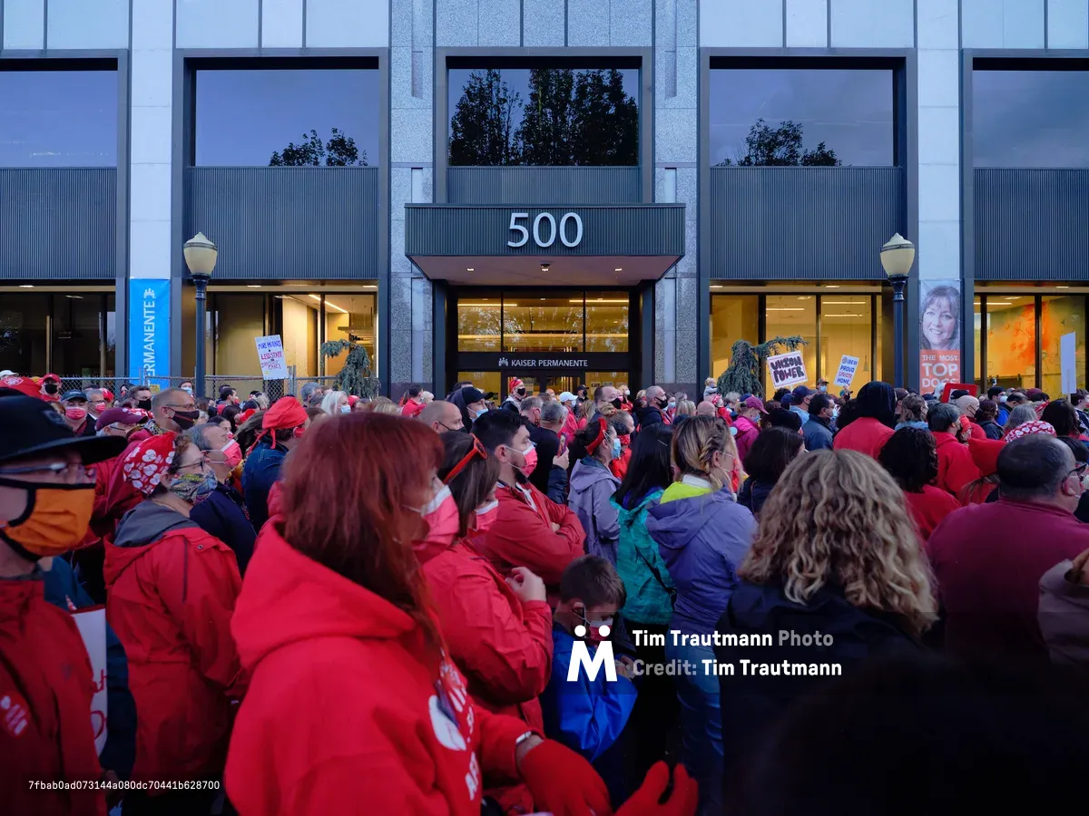 Hundreds of nurses in vibrant red union jackets and winter coats converge in the twilight hours outside the Kaiser Permanente Tower on Northeast Multnomah Street in Portland's Lloyd District. The crowd creates a sea of crimson against the modern glass facade of the 500 building, their collective presence illuminated by the warm glow spilling from the medical facility's entrance. The atmospheric blue hour lighting captures the gravity and determination of healthcare workers preparing for labor action, with protest signs visible among the densely packed crowd.