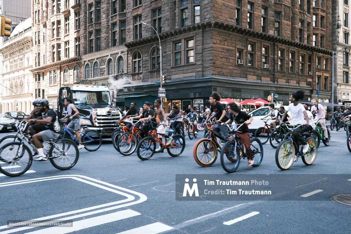 A dynamic river of cyclists flows through the intersection at 7th Avenue in Midtown Manhattan, their colorful bicycles creating streaks of motion against the backdrop of historic brownstone architecture. The late afternoon light bathes the weathered facades in warm golden tones while participants in what appears to be a Critical Mass ride reclaim the urban streetscape. Motion blur captures the kinetic energy of dozens of riders as they navigate the intersection, transforming the typically car-dominated thoroughfare into a temporary cycling sanctuary.