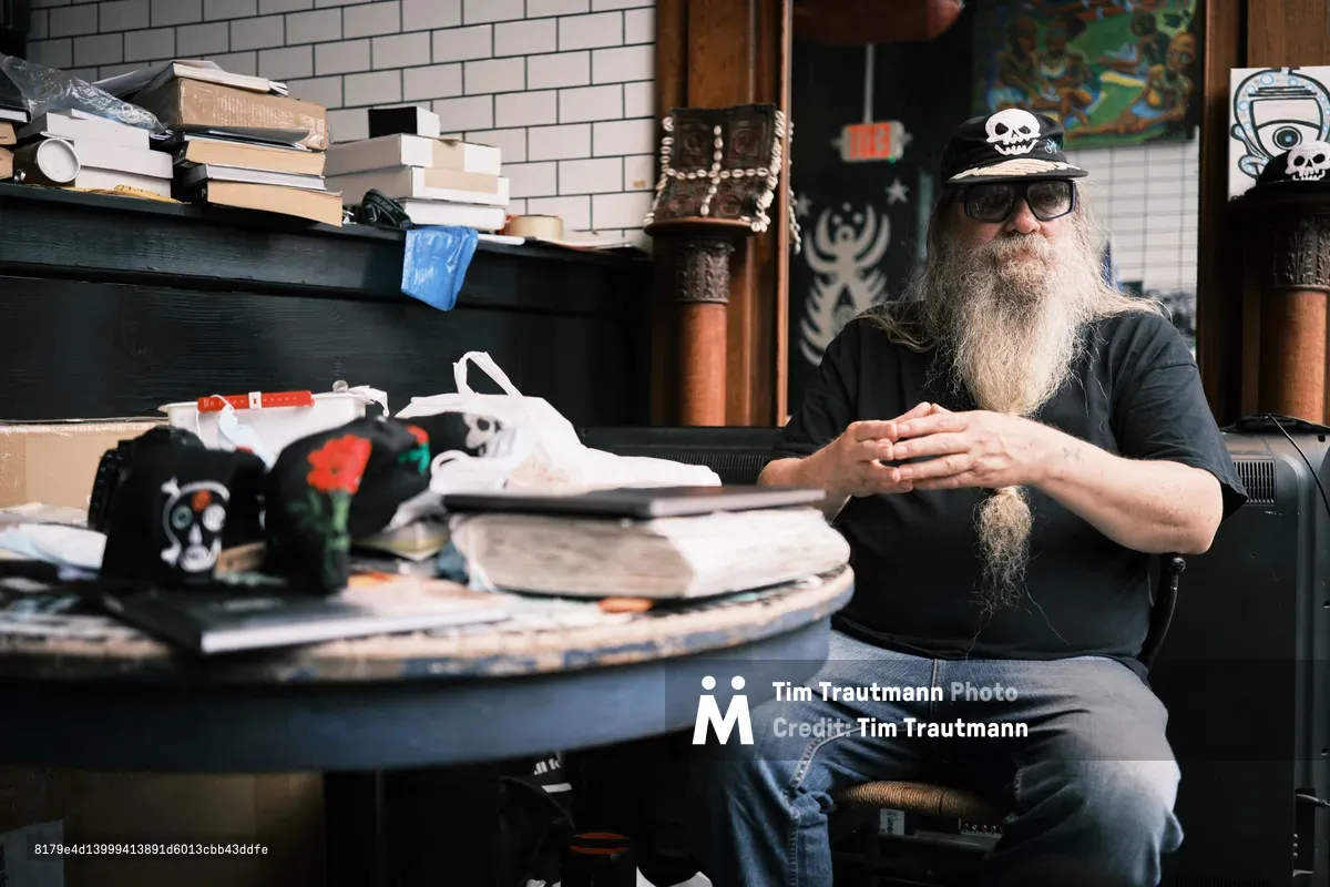 Folk historian and documentarian Clayton Patterson sits contemplatively in his cluttered Essex Street studio, his distinctive long gray beard and skull-adorned cap reflecting decades of chronicling Manhattan's Lower East Side. Warm afternoon light filters through the space, illuminating stacks of books, scattered baseball caps with skull motifs, and the accumulated detritus of a life spent preserving neighborhood history. His weathered hands rest thoughtfully as he surveys his domain—part archive, part sanctuary—where the counter-cultural spirit of old New York endures.