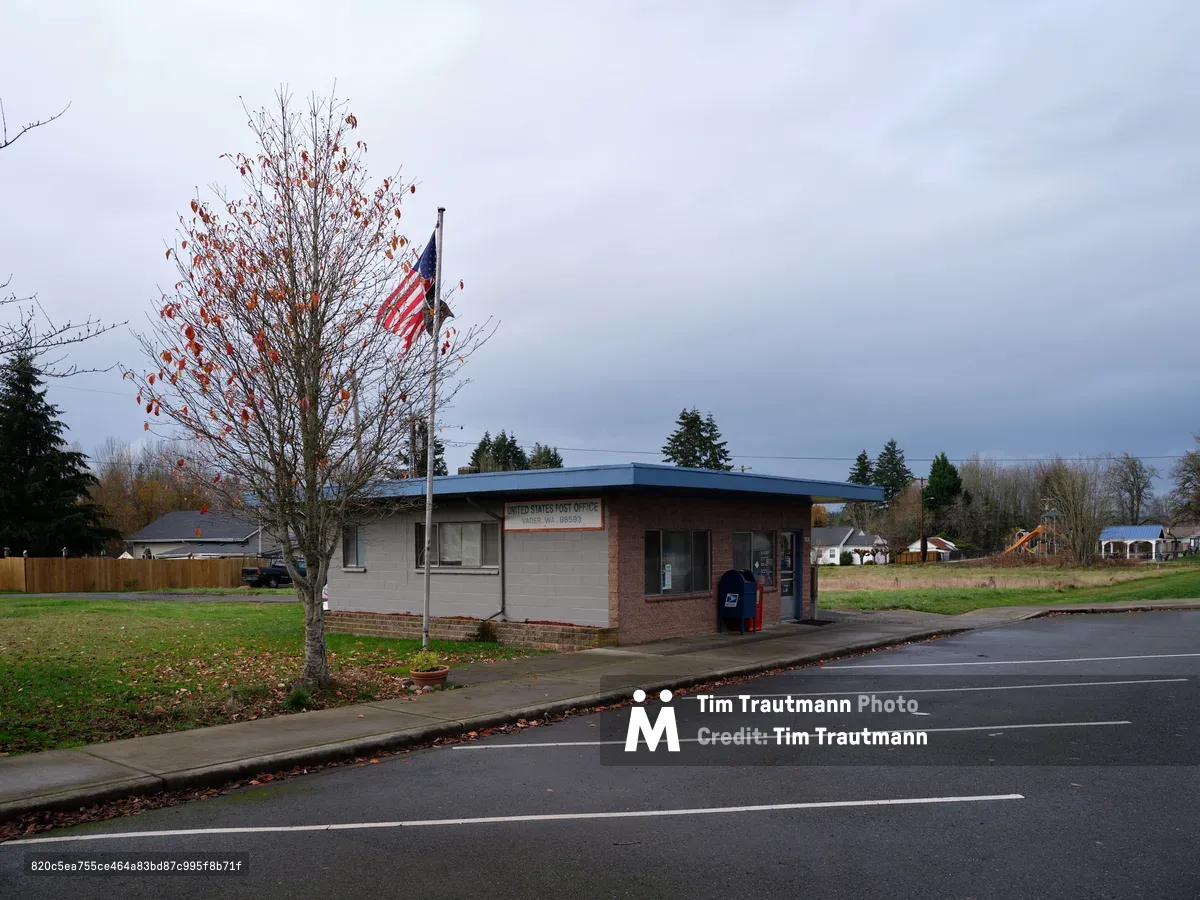 A modest United States Post Office stands sentinel along a quiet street in Vader, Washington, its brick and concrete facade weathered by Pacific Northwest seasons. The American flag hangs motionless against an overcast sky, while a nearby deciduous tree displays autumn's russet leaves, some scattered across the wet pavement. The scene captures the enduring presence of civic infrastructure in rural America, where utilitarian architecture serves communities with understated dignity beneath brooding gray clouds.