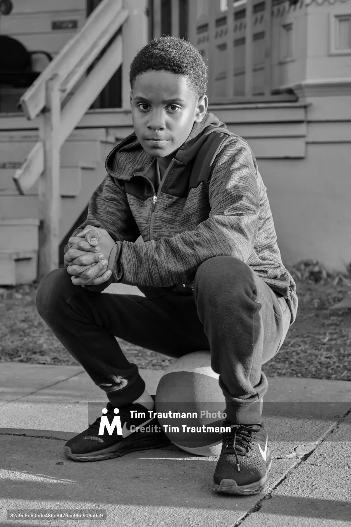 A black-and-white portrait of Kaye, a young boy, crouching on a sidewalk on Mississippi Avenue in Portland, Oregon. He wears a zip-up hoodie, sweatpants, and Nike sneakers, with his hands clasped together and resting on his knees. A basketball sits beneath his feet. He gazes directly into the camera with a steady, composed expression. A wooden porch and the facade of a residential house are visible in the background.