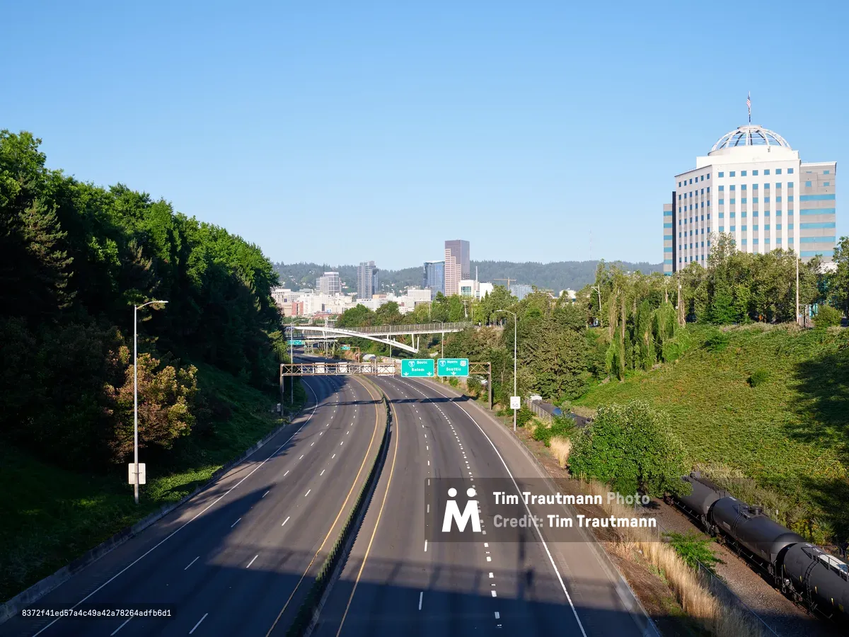 A multi-lane highway curves toward the downtown Portland, Oregon skyline, featuring modern office buildings and skyscrapers surrounded by lush green hills. The road is flanked by dense forests and includes green highway signs directing traffic toward the city center.