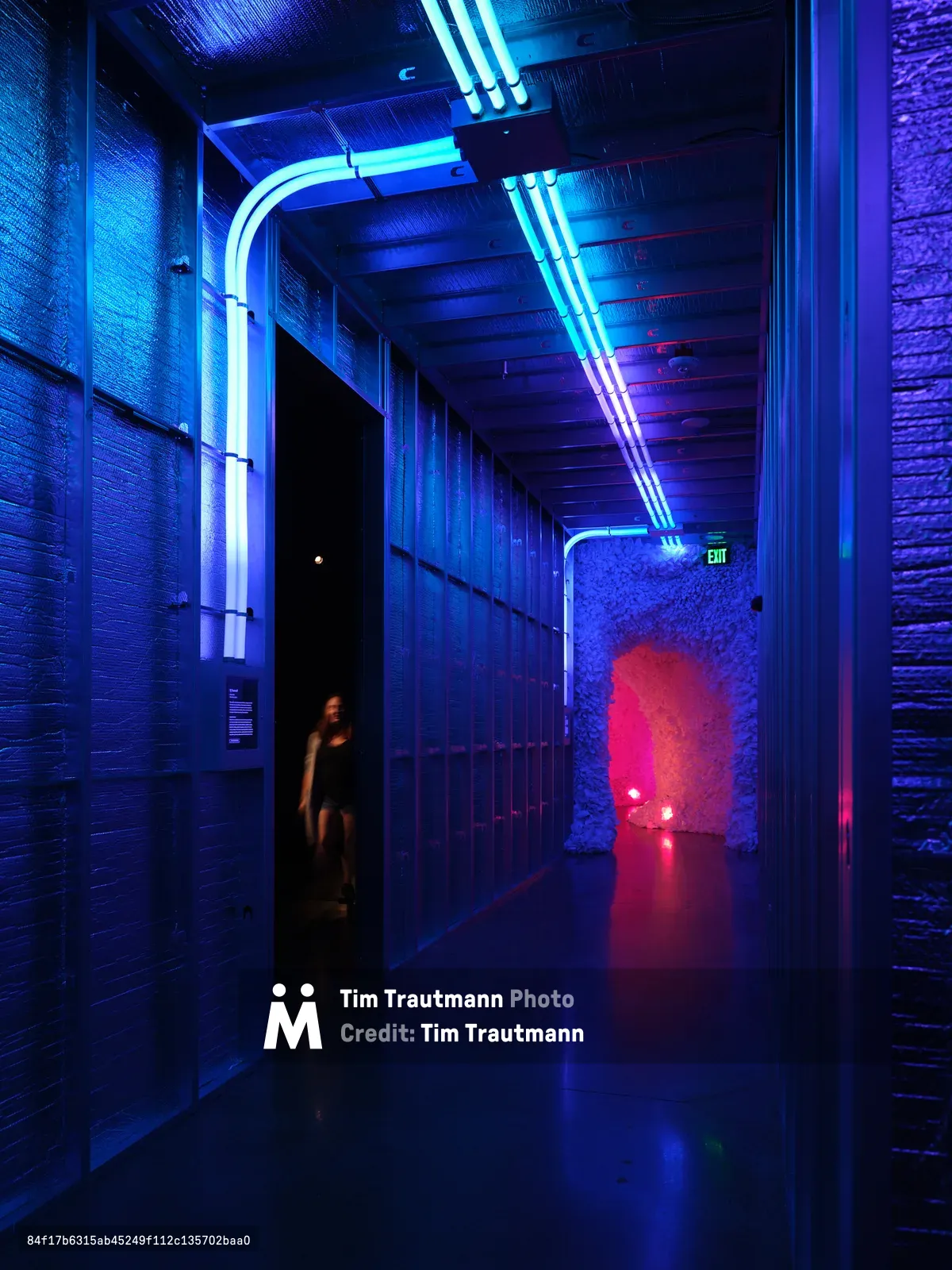 A dramatic industrial hallway illuminated by bright blue and cyan neon lighting, with exposed brick walls and ceiling beams creating a cyberpunk atmosphere. A silhouetted figure stands in a doorway while warm red light glows from an alcove at the corridor's end.