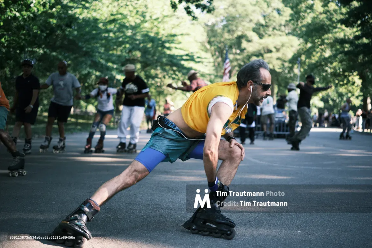 A man in a vibrant yellow tank top and teal shorts leans into a graceful skating stride along Central Park's sun-dappled pathways. The afternoon light filters through the dense canopy above, creating a mosaic of shadows on the asphalt as other skaters and park visitors blur into the verdant background. His focused expression and fluid movement capture the meditative rhythm of urban recreation, while the American flag glimpsed among the trees anchors this quintessentially New York moment.