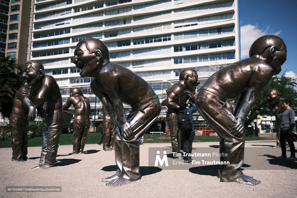 Monumental bronze figures frozen in exuberant laughter populate a sun-drenched plaza in Vancouver's Davie Village, their polished surfaces gleaming against the geometric backdrop of modern residential towers. The sculptural installation captures pure joy in metal, with each larger-than-life figure caught mid-guffaw, heads thrown back in uninhibited mirth. Visitors weave between the sculptures under brilliant blue skies, dwarfed by these towering embodiments of human emotion cast in perpetuity.