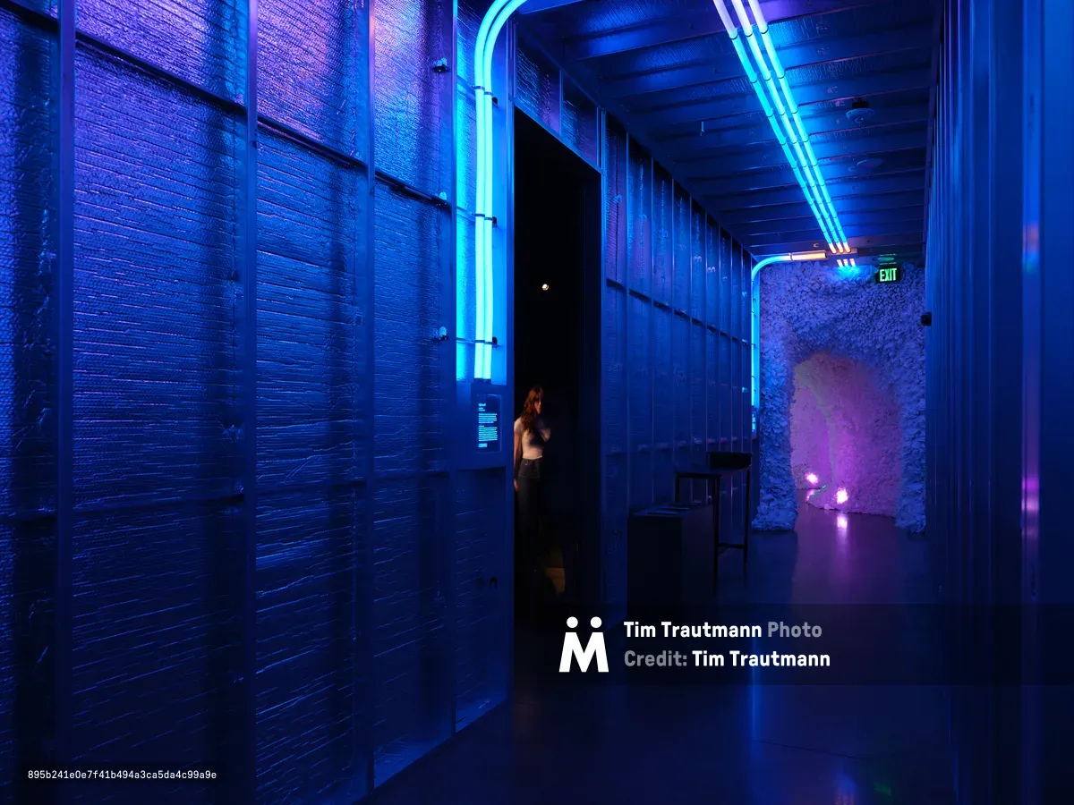 A dramatic blue-lit corridor in Hopscotch, Portland, featuring exposed brick walls, neon lighting, and a person silhouetted in a doorway. The industrial space combines modern lighting design with historic architecture.