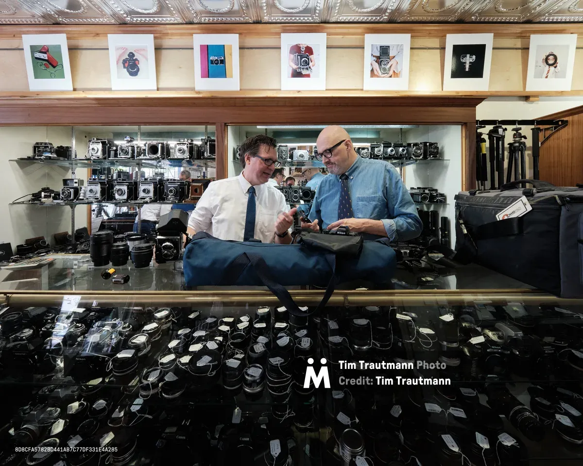 Mike Knight and Arthur Ruckle examine vintage cameras amid the organized chaos of Blue Moon Camera & Machine's Portland storefront. Warm tungsten light bathes the ornate tin ceiling and illuminates rows of meticulously arranged vintage cameras displayed on glass shelves, while hundreds of lenses carpet the floor below. The two specialists lean over their inspection table in focused collaboration, surrounded by decades of photographic history in this legendary Pearl District camera shop.