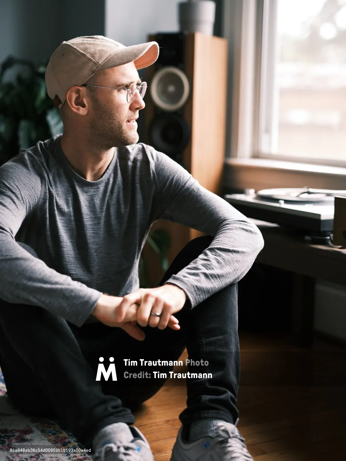 A young man wearing glasses, a baseball cap, and gray long-sleeved shirt sits on the floor of a Richmond neighborhood home in Portland, Oregon, gazing thoughtfully out a bright window. The cozy interior features hardwood floors, a turntable setup, and warm natural lighting.