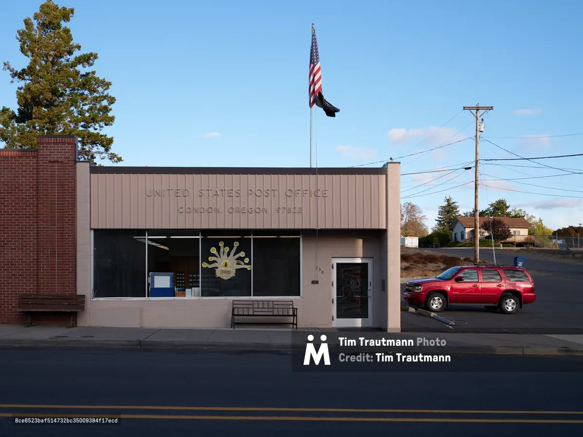 Golden hour light bathes the modest United States Post Office in Condon, Oregon, where mid-century institutional architecture meets small-town practicality. The beige corrugated metal facade displays the building's federal purpose in simple block letters, while a red Chevrolet SUV sits sentinel in the adjacent parking lot beneath a proud American flag. Power lines slice through the crystalline blue sky above this essential community hub, where autumn trees frame the scene with seasonal warmth in Oregon's rural Gilliam County.
