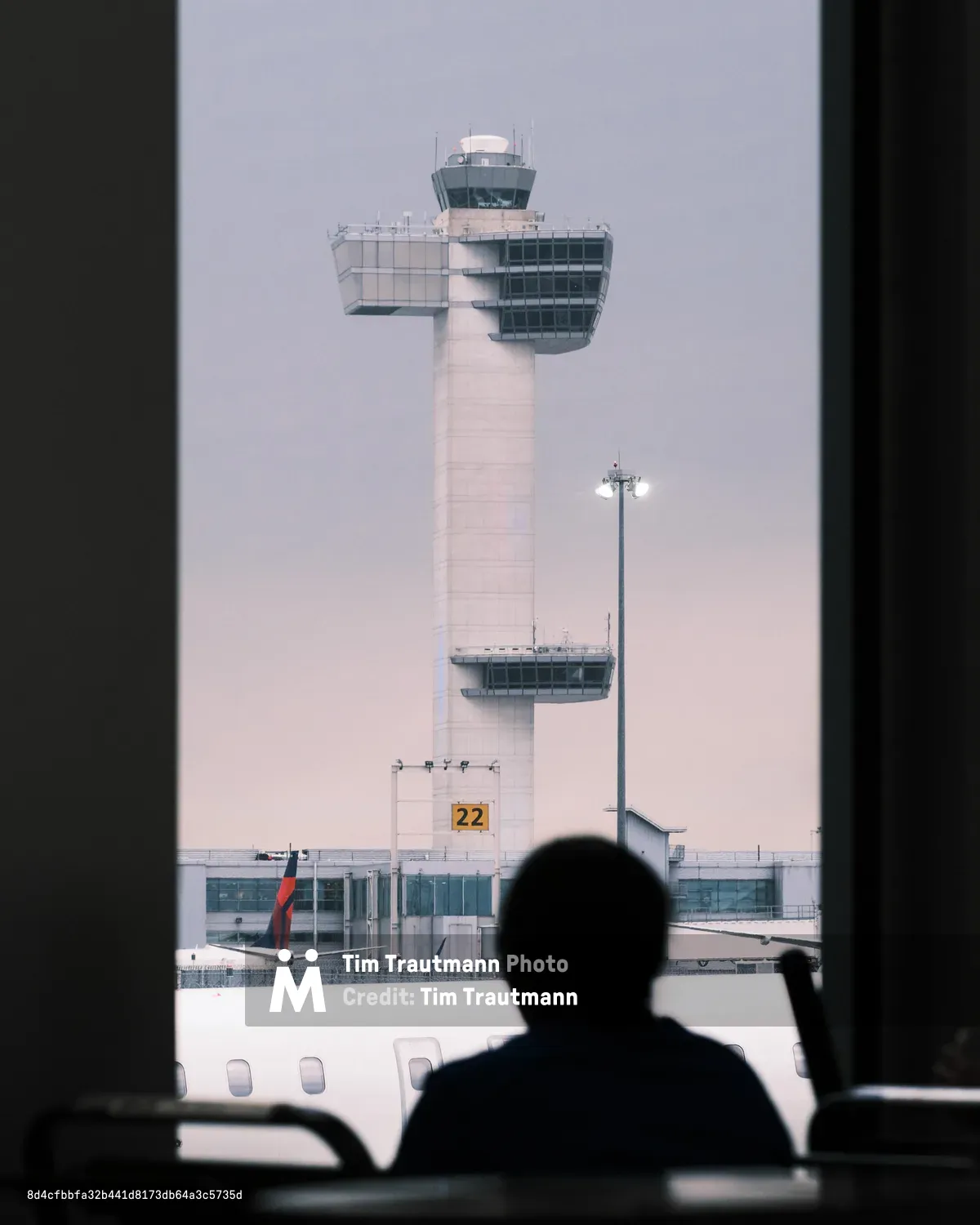 A silhouetted passenger gazes through an airport window at the iconic white control tower of John F. Kennedy International Airport, its distinctive cylindrical form crowned with a glass-enclosed observation deck. The scene captures the liminal moment of air travel, framed by the aircraft's interior in deep shadow against the tower's stark modernist architecture. Soft, diffused light creates an atmospheric haze around the control tower, while gate 22 is visible at its base, anchoring this quintessential airport tableau in Queens.