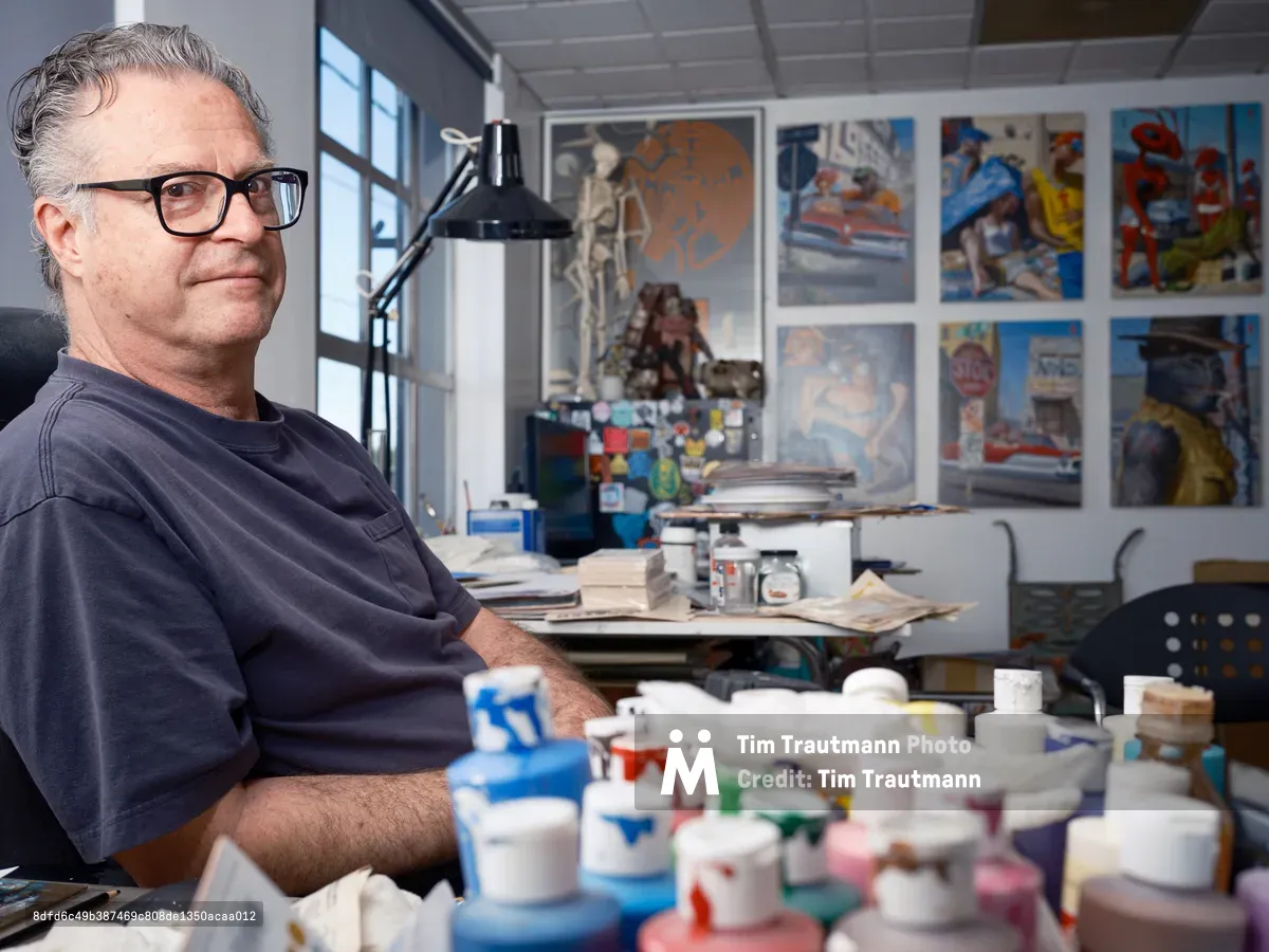 A middle-aged man with glasses and gray hair sits in his art studio surrounded by colorful paint tubes, brushes, and artwork displayed on the walls. The bright, organized workspace features large windows, a desk lamp, and various artistic materials scattered throughout the creative environment.