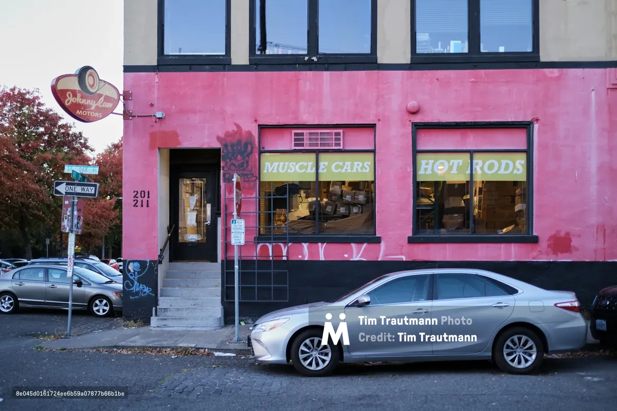 A vibrant magenta-painted automotive shop anchors a Portland street corner, its weathered facade bearing the nostalgic Johnny Law Motors sign and bold yellow window lettering advertising "Muscle Cars" and "Hot Rods." A silver sedan sits parked along the cobblestone street, while autumn foliage frames the scene under crisp blue skies. The building's industrial character speaks to Portland's Central Eastside automotive heritage, where working garages blend seamlessly with the neighborhood's evolving urban landscape.