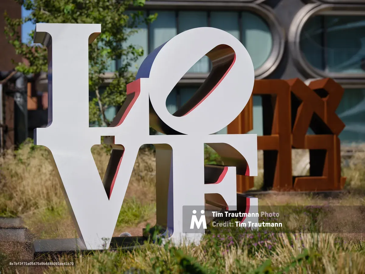 Robert Indiana's iconic LOVE sculpture stands prominently in white lettering against the urban landscape of Manhattan's High Line elevated park. The weathered Cor-Ten steel letters spelling "HOPE" emerge from wild grasses in the background, creating a poetic dialogue between hope and love. Dappled sunlight filters through the scene, illuminating the sculptural forms while the geometric windows of surrounding buildings provide an architectural backdrop. The natural plantings of the linear park soften the industrial heritage of this transformed railway corridor.