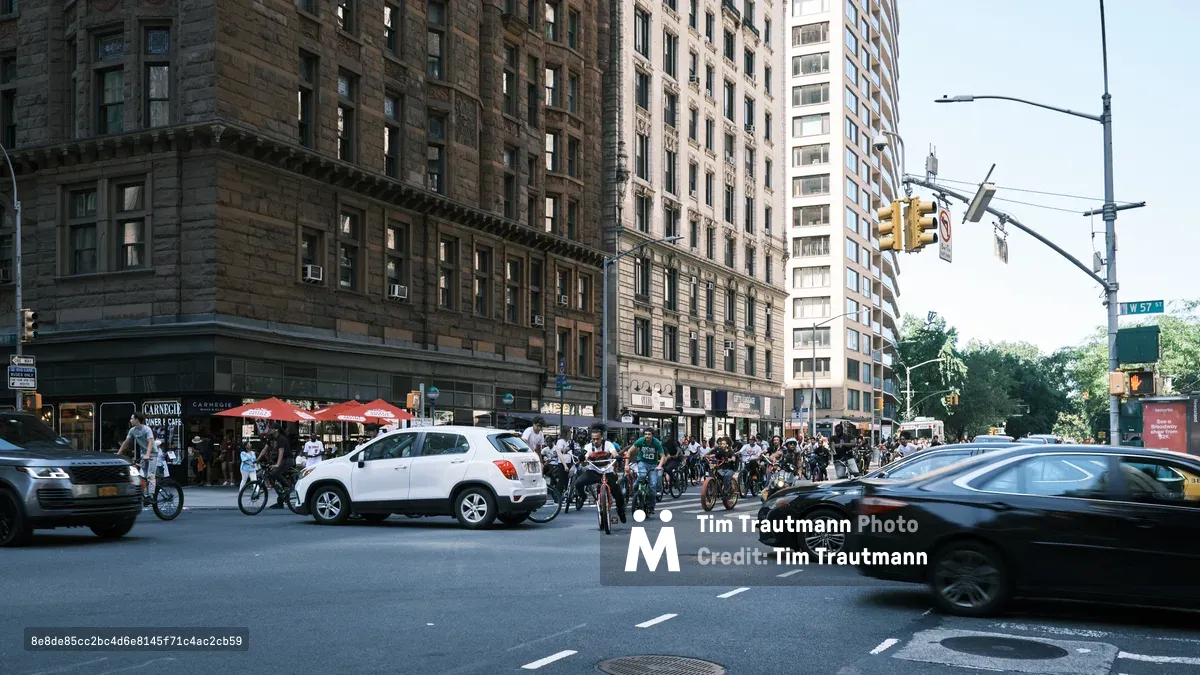 A sea of bicycles clusters at the intersection of West 57th Street and Seventh Avenue in Midtown Manhattan, where riders pause amid the urban choreography of yellow traffic lights and pre-war architecture. The warm afternoon light bathes the weathered brownstone facades and Carnegie Diner's red umbrellas, while modern vehicles blur past in the foreground. This moment captures the tension between old and new New York, where cycling culture claims its space among the towering monuments to commerce and history.