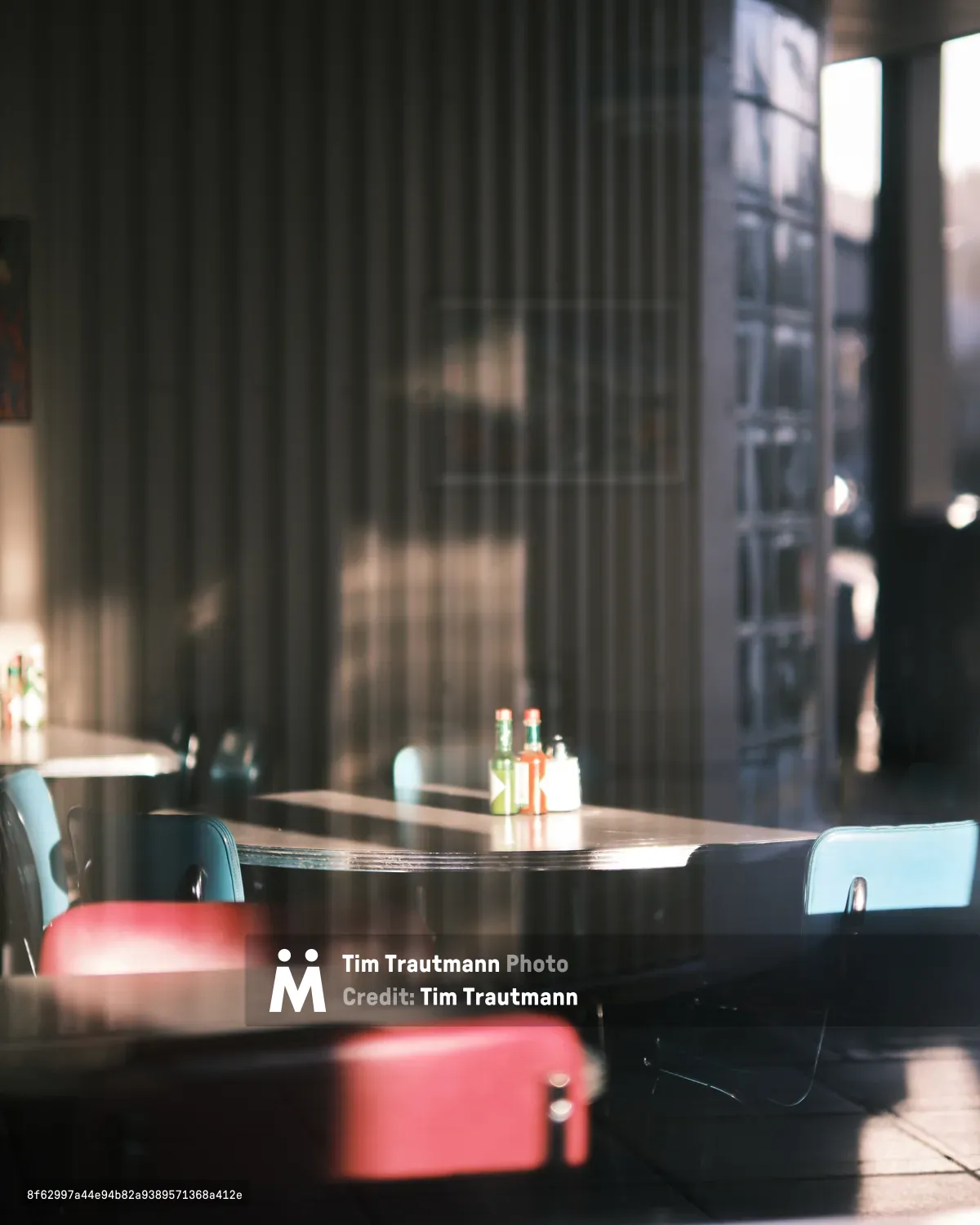 Soft morning light filters through tall windows, casting gentle shadows across a quiet corner table in a Seattle café. The scene captures the intimate atmosphere of urban breakfast culture, with condiment bottles centered on a round wooden table surrounded by red vinyl stools and light blue chairs. The shallow depth of field creates a dreamy quality, with the background dissolving into warm bokeh while the foreground maintains sharp detail on the simple table setting.