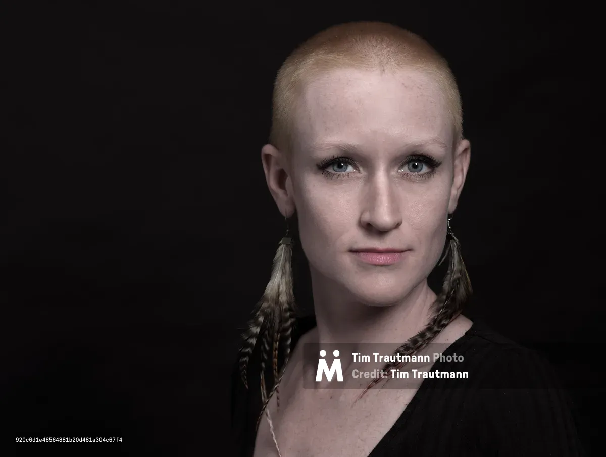Cayce Ryan gazes directly at the camera with quiet intensity, her closely-cropped hair catching the controlled studio lighting against the deep black backdrop. Feathered earrings frame her face, adding organic texture to the minimalist composition, while her subtle makeup and black garment create an understated elegance. The professional headshot, captured in Portland's Factor Building, reveals both vulnerability and strength through precise lighting that sculpts her features with dramatic shadow and highlight.