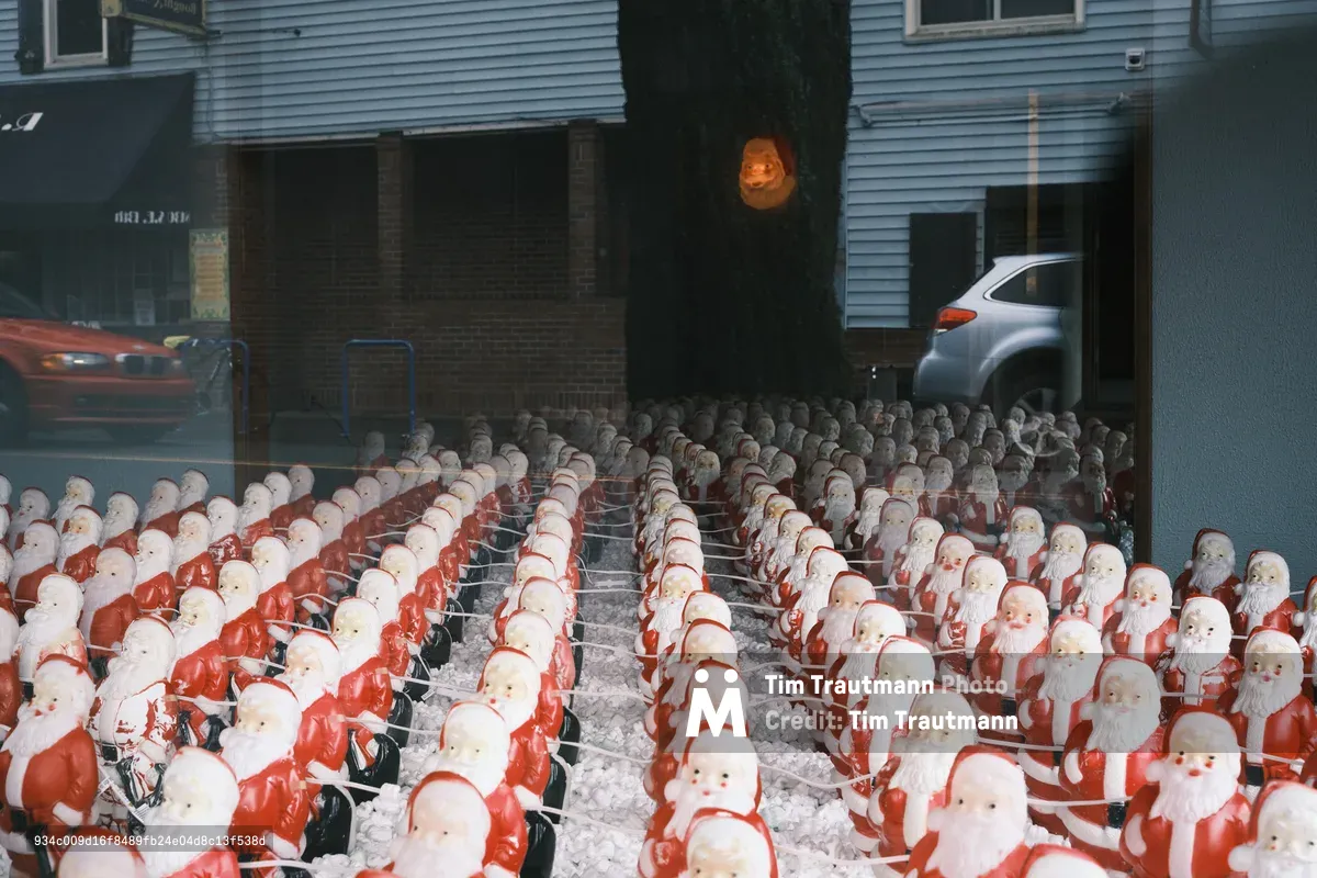 Hundreds of identical Santa Claus figures occupy the floor of Consignment Store in Portland's Sellwood neighborhood, creating Chris Willis's surreal annual art installation. The ceramic or plastic Santas are arranged in precise rows on wire shelving, their jolly faces creating an unsettling sea of mass-produced Christmas cheer. Gallery windows reflect the quiet street outside, where a silver SUV and storefronts hint at the ordinary world beyond this dreamlike tableau of holiday multiplication.