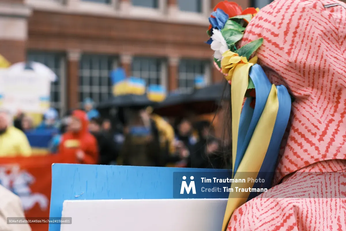 A woman in a coral-striped headscarf adorned with flowing blue and yellow ribbons—the colors of Ukraine—stands at a protest vigil outside Portland's Revolution Hall. The patriotic ribbons cascade from a crown of artificial flowers, creating a striking visual metaphor of hope and defiance. Behind her, blurred demonstrators gather in the urban streetscape, their forms dissolving into the shallow depth of field that isolates this singular moment of quiet determination.