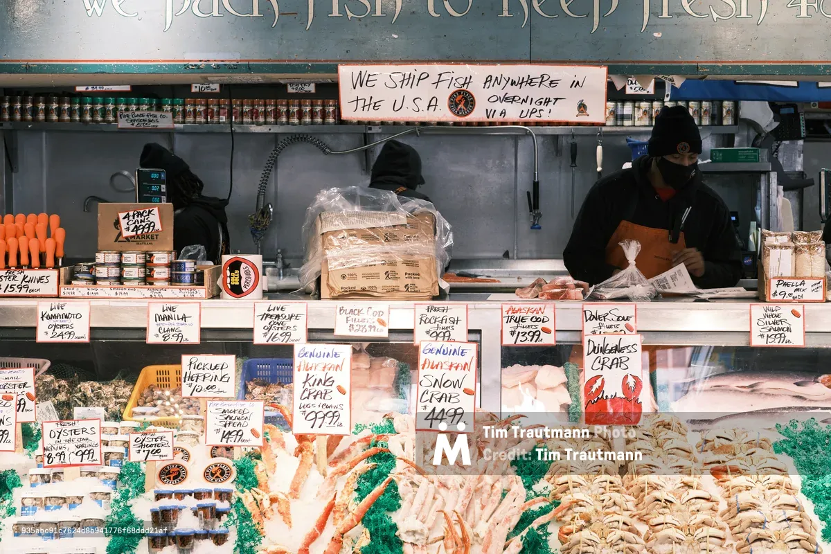 Behind the iconic seafood counter at Seattle's Pike Place Fish Market, workers in black hoodies tend to their daily catch beneath handwritten price signs and shipping notices. The bustling display showcases an abundance of fresh seafood arranged on crushed ice, from Dungeness crab to Alaskan true cod, while bright orange price tags punctuate the scene. The industrial stainless steel workspace and vintage signage create an authentic portrait of this legendary Seattle marketplace, where maritime tradition meets urban commerce.