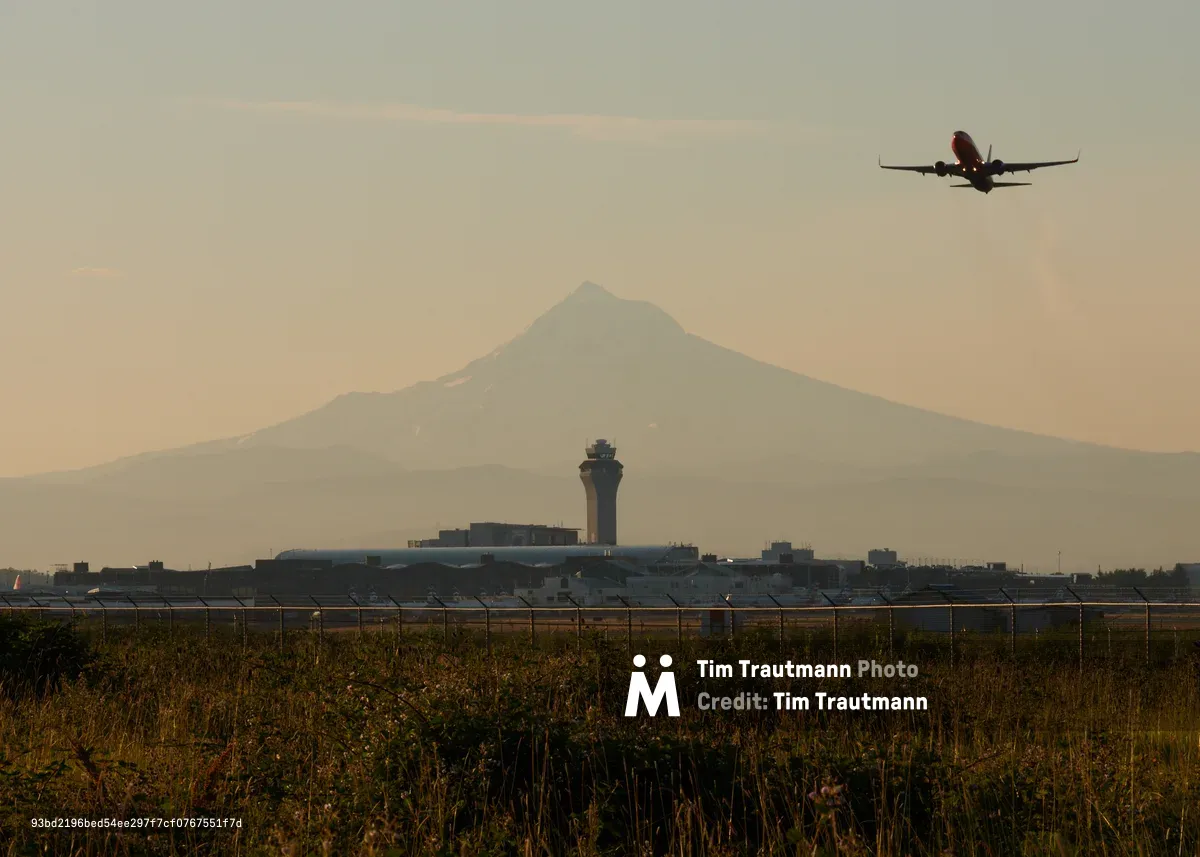 A commercial airliner climbs into the amber-hued twilight sky above Portland International Airport, silhouetted against the majestic volcanic cone of Mount Hood. The airport's distinctive control tower stands sentinel in the middle distance, while wild grasses in the foreground frame this layered composition of human aviation intersecting with Oregon's iconic Cascade peak. The warm, diffused light suggests either early morning or evening departure, casting the entire scene in a nostalgic, golden atmosphere.