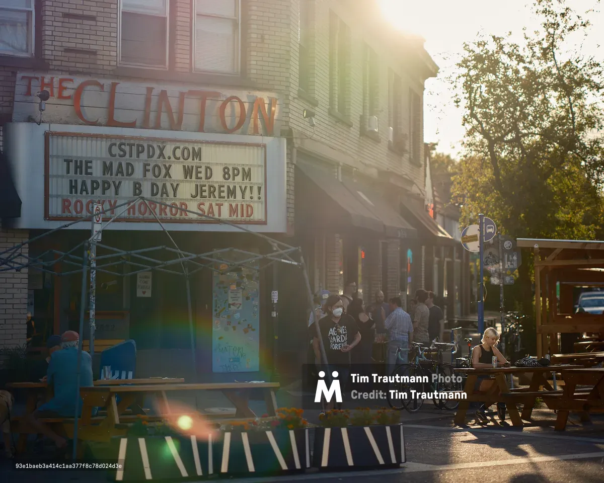 Alt text:  The exterior of The Clinton Street Theater in Portland, Oregon, bathed in warm golden lens-flare sunlight. The theater's marquee reads "The Mad Fox Wed 8PM — Happy B Day Jeremy!! — Rocky Horror Sat Mid." A lively crowd of people gathers on the sidewalk outside, with outdoor picnic tables and planters with marigolds occupying a parklet in the foreground. A woman in a face mask wearing a black "Agnès Varda Forever" t-shirt walks past. Bicycles are parked along the curb, and a woman sits alone at a picnic table to the right.
