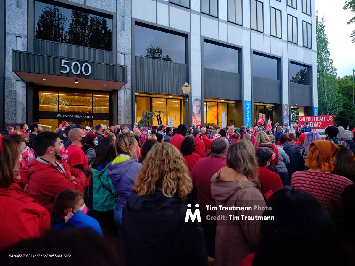 A sea of red-clad healthcare workers fills the sidewalk outside Kaiser Permanente's modernist headquarters at 500 Northeast Multnomah Street in Portland's Lloyd District. The crowd of striking nurses creates a powerful visual contrast against the building's sleek glass facade and concrete columns, their unified presence illuminated by the warm glow spilling from the medical center's ground-floor windows in the gathering dusk. Signs demanding "CAN YOU HEAR US NOW?" punctuate the peaceful but determined assembly, while the institutional architecture looms overhead, emphasizing the David-versus-Goliath nature of the labor dispute.