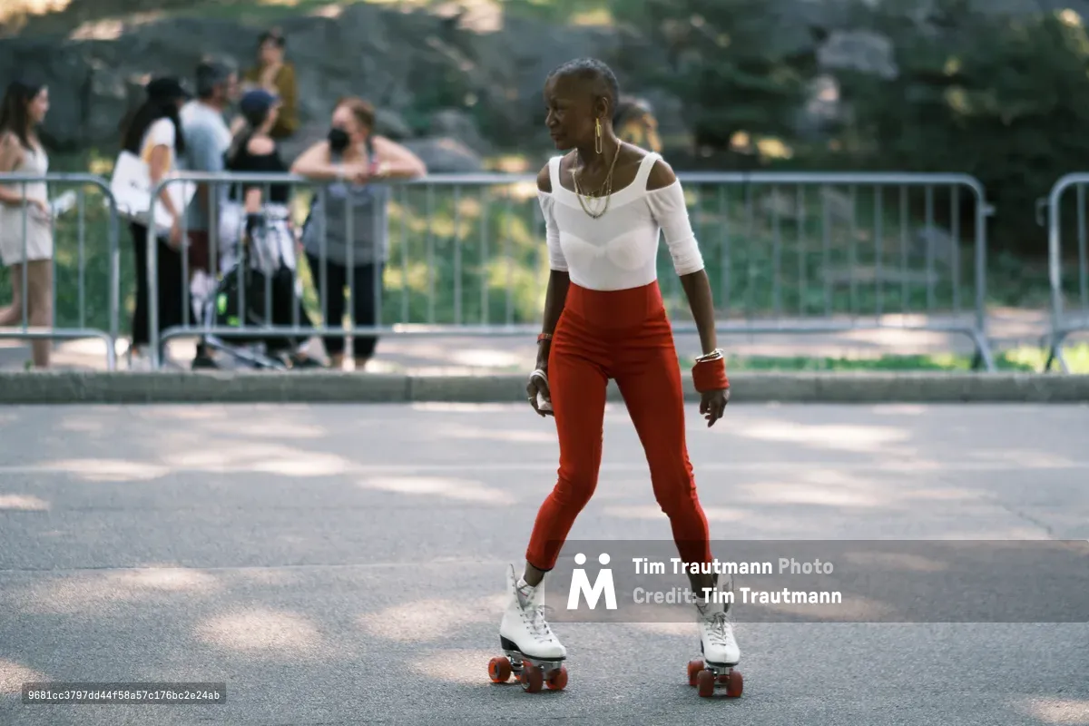 A woman glides gracefully on vintage white roller skates across the sun-dappled asphalt of Central Park's skating rink. She wears a striking ensemble of vermillion high-waisted pants and a crisp white cold-shoulder top, her movements captured mid-stride as golden hour light filters through the leafy canopy above. Behind her, metal barriers frame other park visitors who blur into the dreamy bokeh of late afternoon shadows and light. The scene pulses with nostalgic energy and urban recreation.
