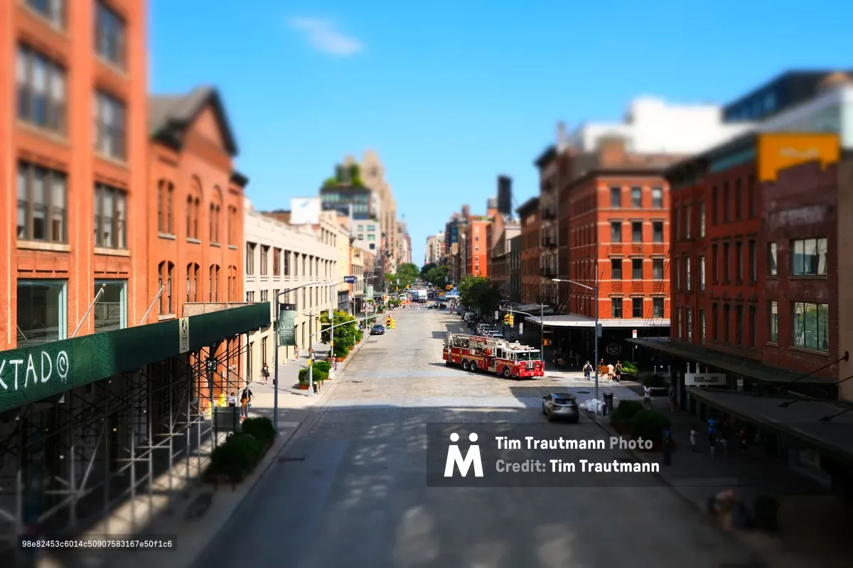 A dramatic tilt-shift perspective captures the intersection of 10th Avenue and West 14th Street from the elevated High Line walkway, where industrial-era brick warehouses frame a sun-drenched corridor. A red fire truck commands the center of the composition, its emergency presence juxtaposed against the leisurely pedestrians exploring this transformed Meatpacking District landscape. The shallow depth of field renders the urban canyon in miniature-like detail, while brilliant blue skies crown the historic neighborhood's rebirth from industrial grit to cultural destination.