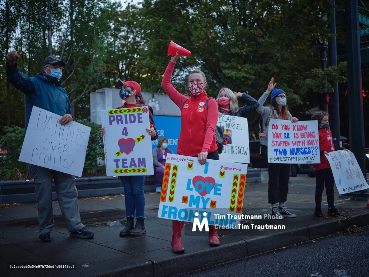Healthcare workers gather on the sidewalk outside Kaiser Permanente Tower in Portland's Lloyd District, their voices unified in protest against inadequate staffing levels. Masked demonstrators hold colorful handmade signs declaring 'Love My Frontline' and demanding safe patient-to-nurse ratios, their red scrubs and bright placards creating vivid punctuation marks against the muted autumn evening. The golden hour light filters through towering street trees, casting long shadows across the concrete as nurses raise their fists and megaphones in solidarity. The scene captures the determined spirit of frontline workers advocating for both patient safety and workplace dignity during a pivotal labor action.