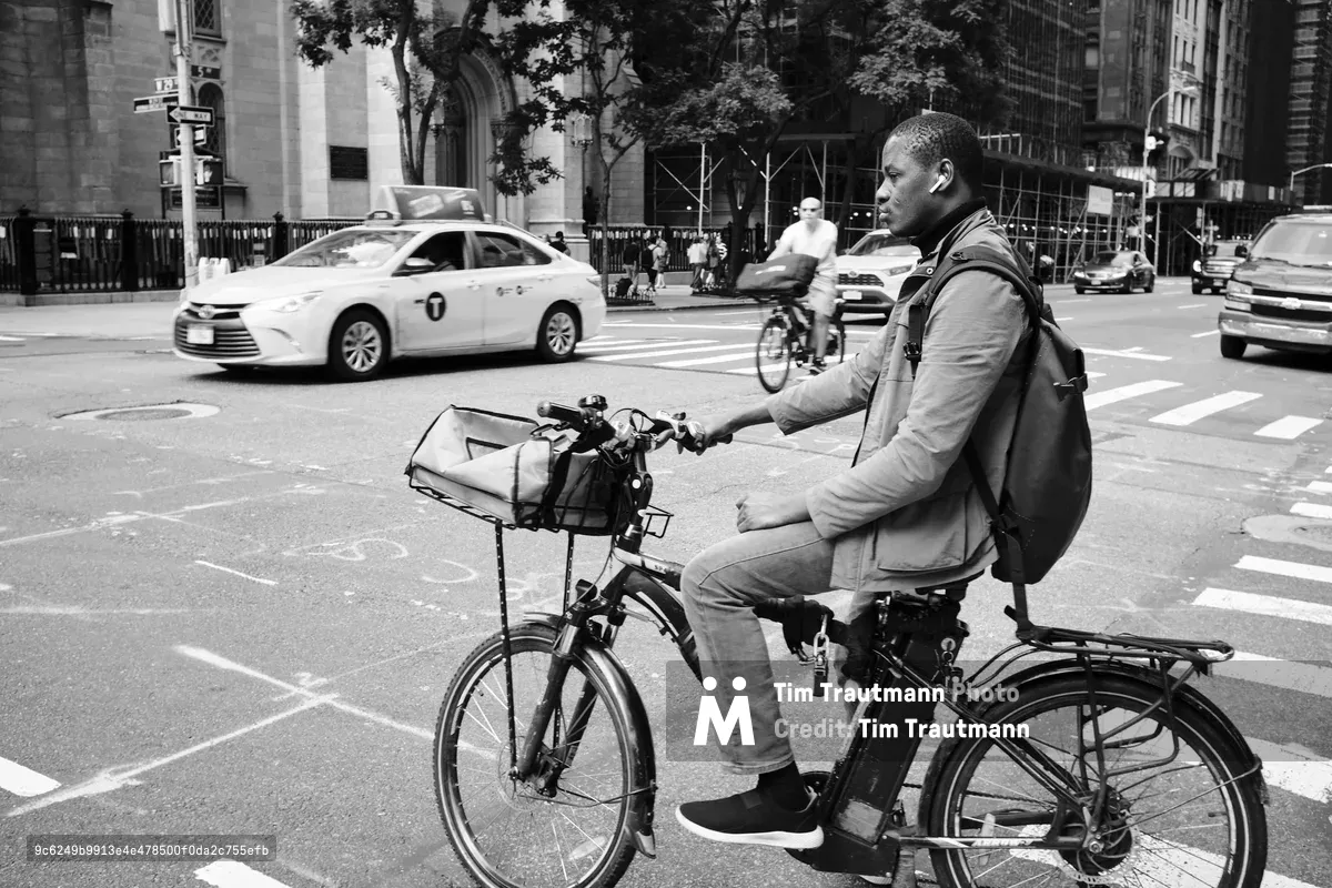 A delivery cyclist pauses at the intersection of East 29th Street in Manhattan's NoMad district, his weathered face caught in contemplative profile against the bustling urban backdrop. The black and white composition captures the gritty authenticity of city life, with his utilitarian bicycle equipped with a front cargo basket positioned prominently in the crosswalk. Behind him, the familiar yellow taxi and Gothic Revival architecture of the neighborhood create a quintessentially New York tableau, while dappled sunlight filters through street trees onto the asphalt.