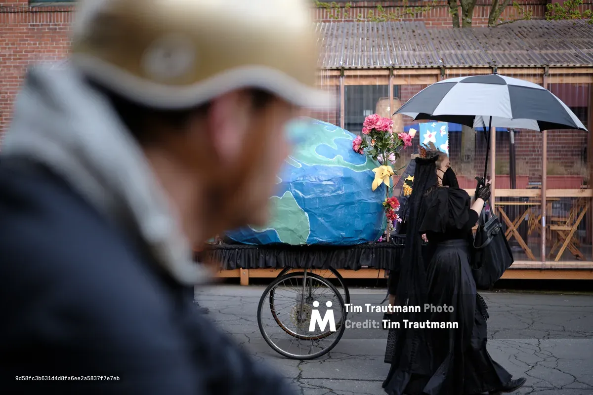 A solemn funeral scene in Portland, Oregon showing mourners in black gathered around an ornate horse-drawn hearse decorated with blue fabric and flowers, with an umbrella providing shelter during what appears to be a rainy day.