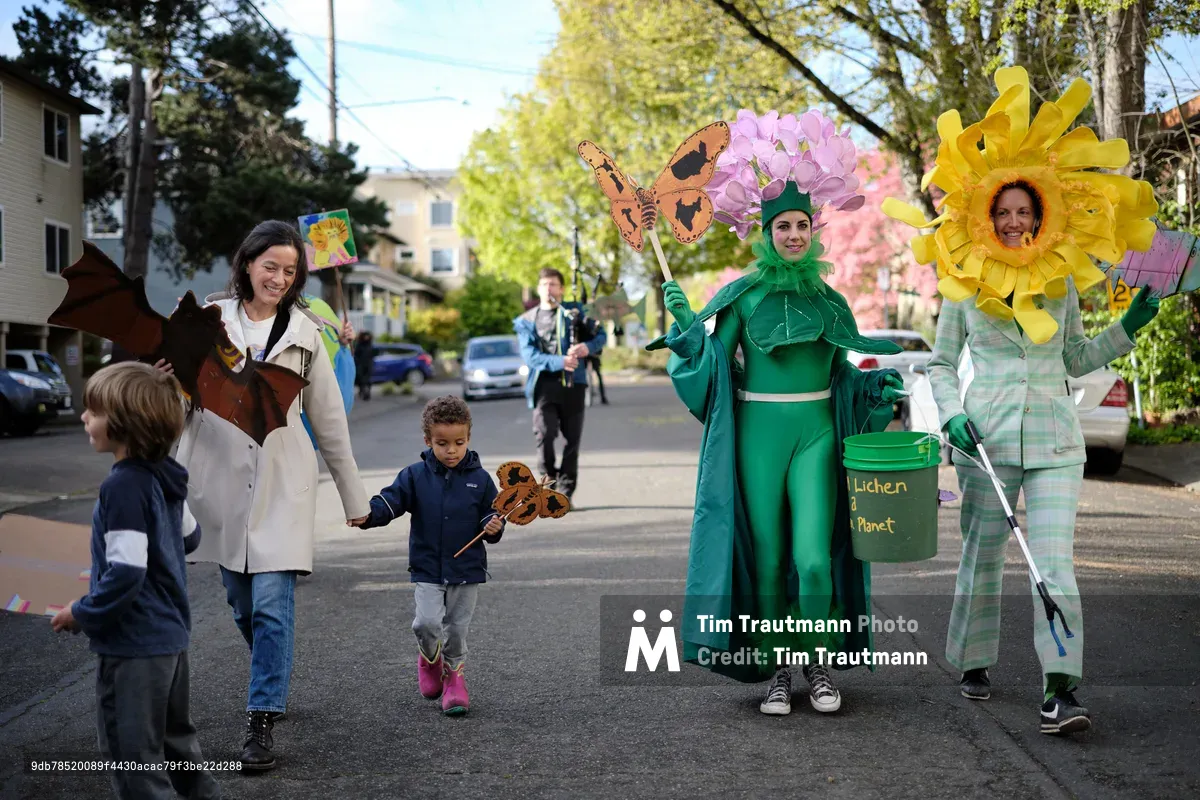 A group of environmental activists and families participate in a colorful street parade in Portland, Oregon, featuring participants dressed as flowers, trees, and nature-themed costumes while carrying eco-friendly signs and props. Children and adults walk together on a residential street lined with trees and houses, promoting environmental awareness through creative costumes and community engagement.
