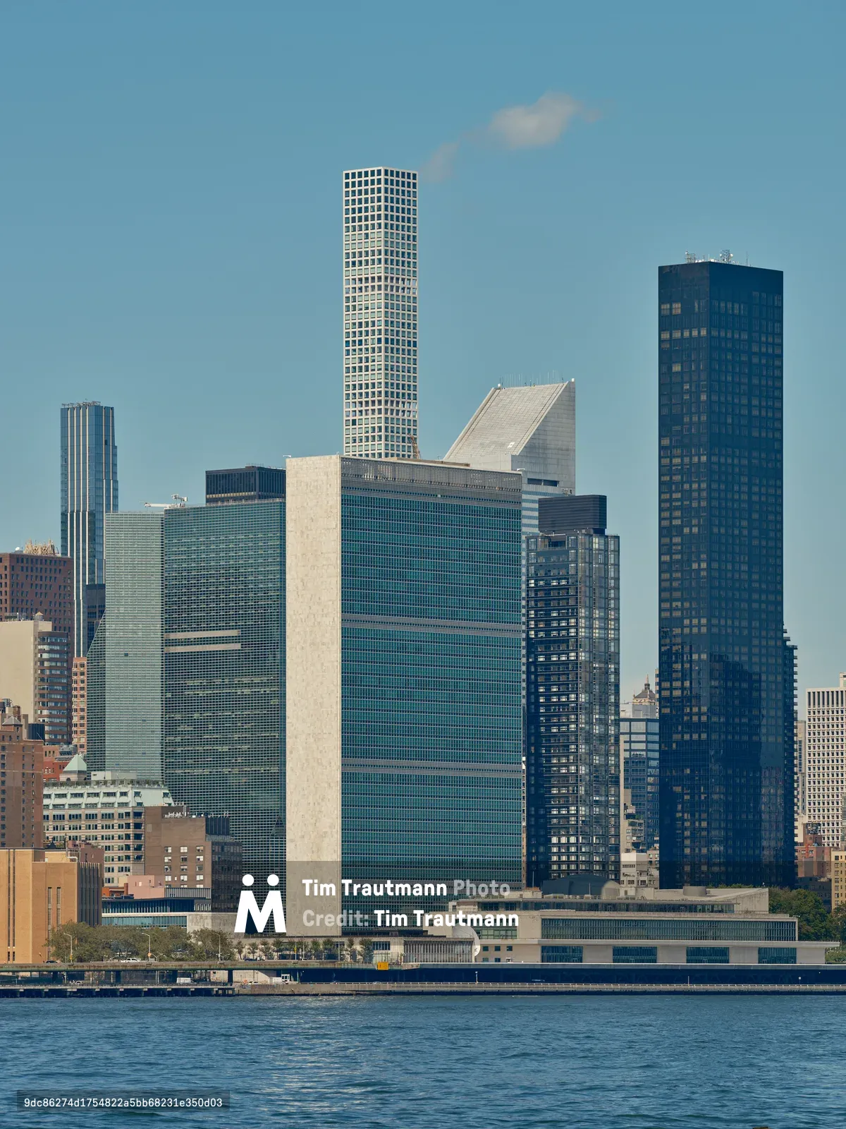 The iconic Manhattan skyline rises majestically from the East River, captured from Brooklyn's Greenpoint neighborhood on a crystalline afternoon. The United Nations Headquarters' distinctive rectangular form anchors the composition alongside gleaming residential towers, their glass facades catching the clear light against an azure sky. Gentle ripples disturb the river's surface in the foreground, while wispy clouds drift across the expansive urban panorama. The interplay of modernist architecture and classical skyscraper forms creates a compelling study of New York's evolving vertical landscape.