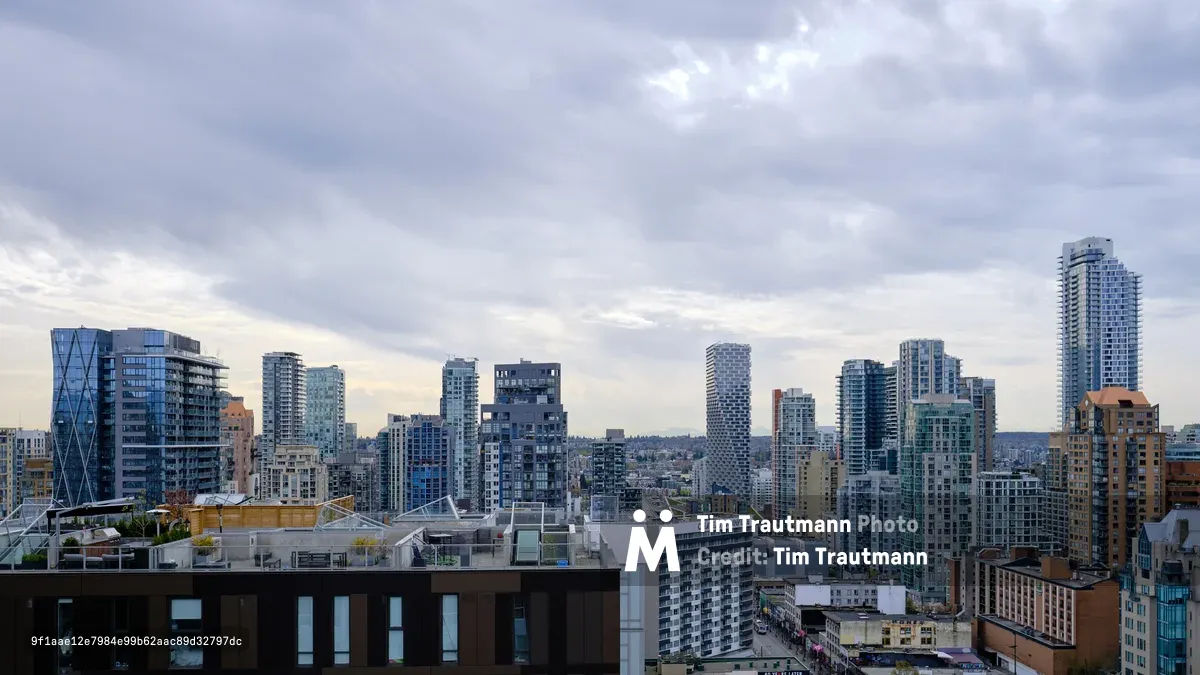 The dense urban fabric of Vancouver's Yaletown and downtown core stretches toward the horizon under a dramatic overcast sky, where pewter clouds create moody illumination across the glass and concrete towers. Modern residential high-rises with distinctive architectural elements—including a prominent twisted tower and geometric facades—punctuate the cityscape, while older brick buildings in the foreground provide textural contrast. The late afternoon light filters through the cloud cover, casting the scene in cool blues and muted tones that emphasize the metropolitan density of this Pacific Northwest metropolis.