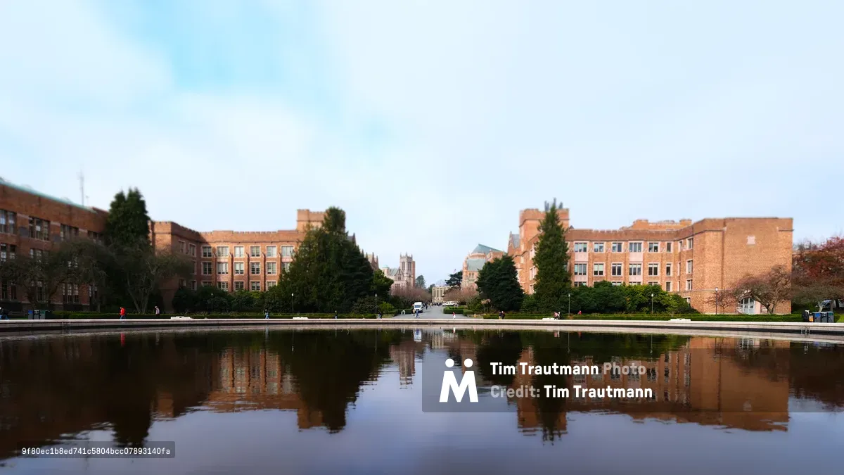 The iconic Drumheller Fountain creates a perfect mirror on the University of Washington's Rainier Vista, doubling the stately Georgian Revival brick dormitories that frame this grand campus axis. Golden hour light warms the red brick facades while towering evergreens punctuate the symmetrical composition. In the distance, the Gothic Revival spires of the campus emerge through the soft atmospheric haze, while students move like distant figures across the manicured grounds.
