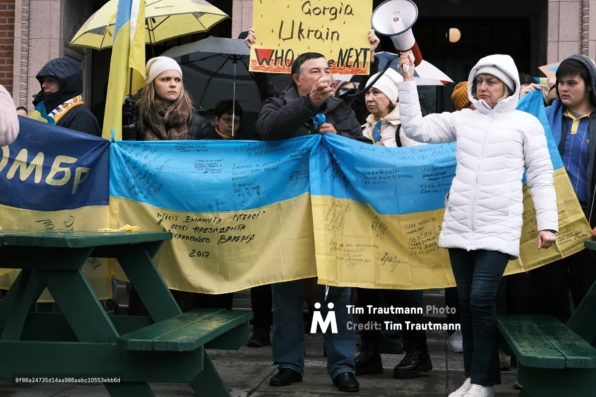 Ukrainian demonstrators gather on the weathered steps of Portland's Revolution Hall, their hands gripping a signature-laden blue and yellow flag that ripples with the weight of collective hope and dread. A man in dark clothing speaks passionately into a microphone while protesters in white winter coats stand resolute behind him, yellow umbrellas blooming overhead like defiant flowers against the overcast Pacific Northwest sky. Signs reading 'Georgia Ukraine WHO is NEXT' pierce through the crowd, their urgent handwriting capturing the prescient anxiety of a community bracing for war.