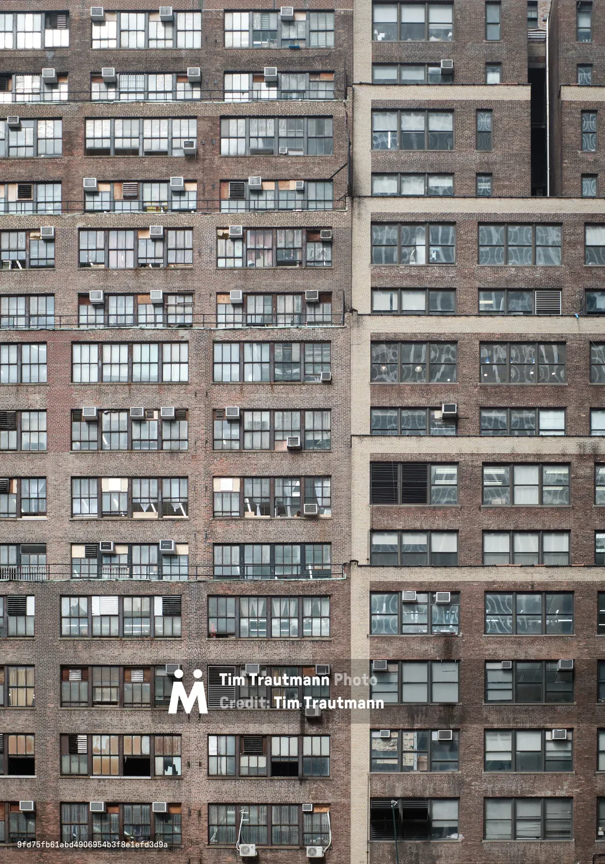 A dense pattern of weathered brick apartment building facades creates an urban tapestry in Midtown Manhattan. The repetitive grid of windows, punctuated by air conditioning units and varying states of wear, reveals the intimate details of city living through glimpses of curtains, blinds, and personal belongings. The warm brown and red brick tones contrast with the cool reflective glass, while the geometric composition emphasizes the vertical density of New York residential architecture.