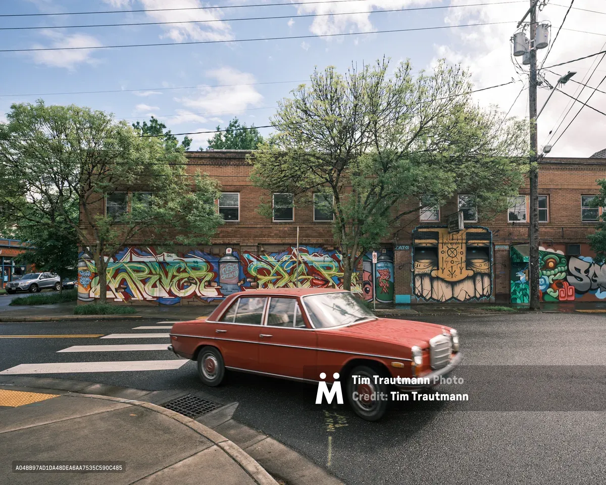 A crimson vintage Mercedes-Benz sedan glides through the intersection at Southeast Division Street in Portland's Richmond neighborhood, its classic lines contrasting against a vibrant wall of street art adorning the former Oregon Theater. The weathered brick building serves as an urban canvas, its colorful graffiti murals creating a dynamic backdrop under the dappled afternoon light filtering through mature street trees. Power lines cross overhead like geometric sketches against the soft blue sky, while the vintage automobile becomes a moving piece of the neighborhood's evolving artistic identity.