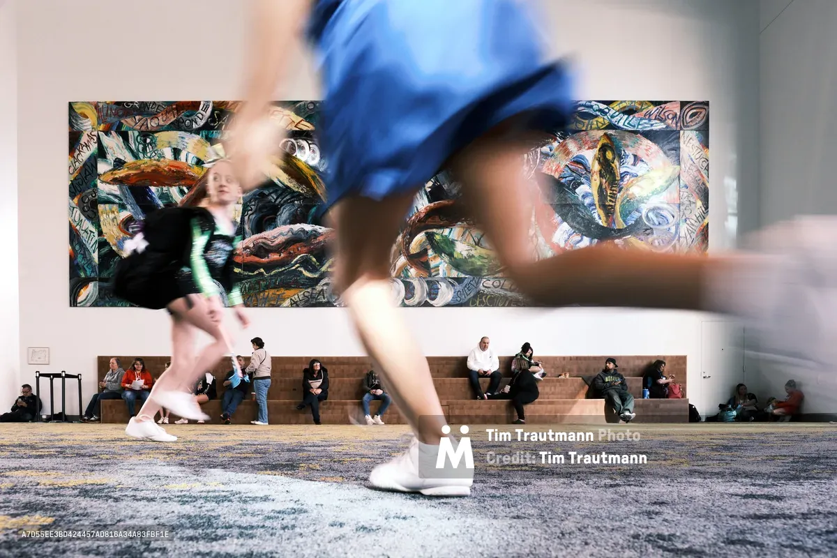 Blurred figures in motion streak across the foreground as visitors hurry through the spacious foyer of Portland's Oregon Convention Center. Behind them, spectators rest on terraced concrete steps beneath a vibrant, large-scale mural featuring serpentine forms and bold street art aesthetics. The juxtaposition of kinetic energy and static contemplation creates a dynamic urban tableau, with warm lighting illuminating the contemporary architectural space.