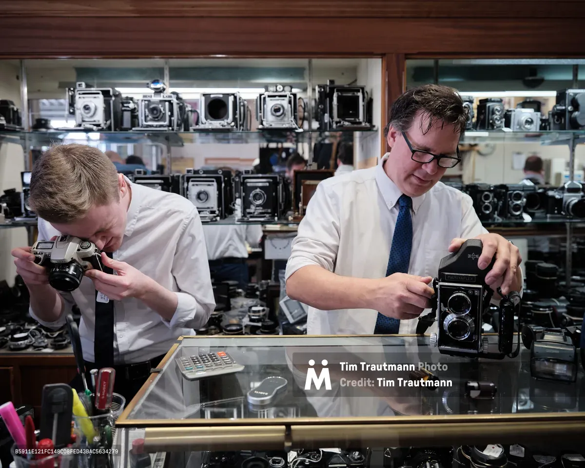 In the intimate confines of Blue Moon Camera & Machine, Portland's legendary camera haven, Mike Knight and a colleague work with reverent concentration behind a gleaming glass counter. The warm tungsten light illuminates their careful examination of vintage equipment while towering displays of classic cameras—from twin-lens reflex models to large format view cameras—create a museum-like backdrop. The scene captures the quiet dedication of analog photography's guardians, their focused gestures speaking to decades of expertise amid this treasure trove of mechanical precision.