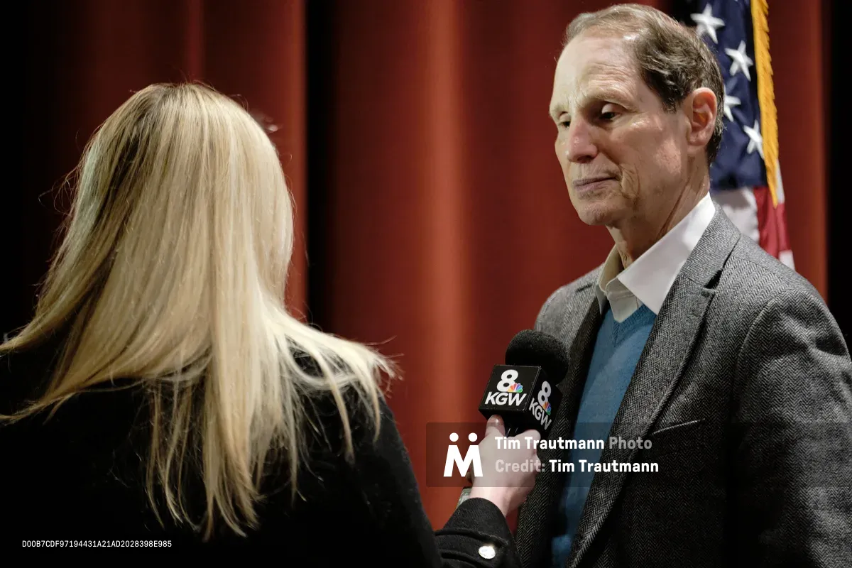 Oregon Senator Ron Wyden stands composed yet contemplative as he addresses questions from a KGW Channel 8 television reporter in what appears to be a government building in Portland's Saint Johns neighborhood. The intimate interview setup captures the senator in a charcoal wool blazer and teal shirt, his weathered features reflecting the gravity of political discourse. Rich burgundy wood paneling and the American flag create a backdrop of institutional gravitas, while the blonde reporter's microphone bridges the space between public servant and press.