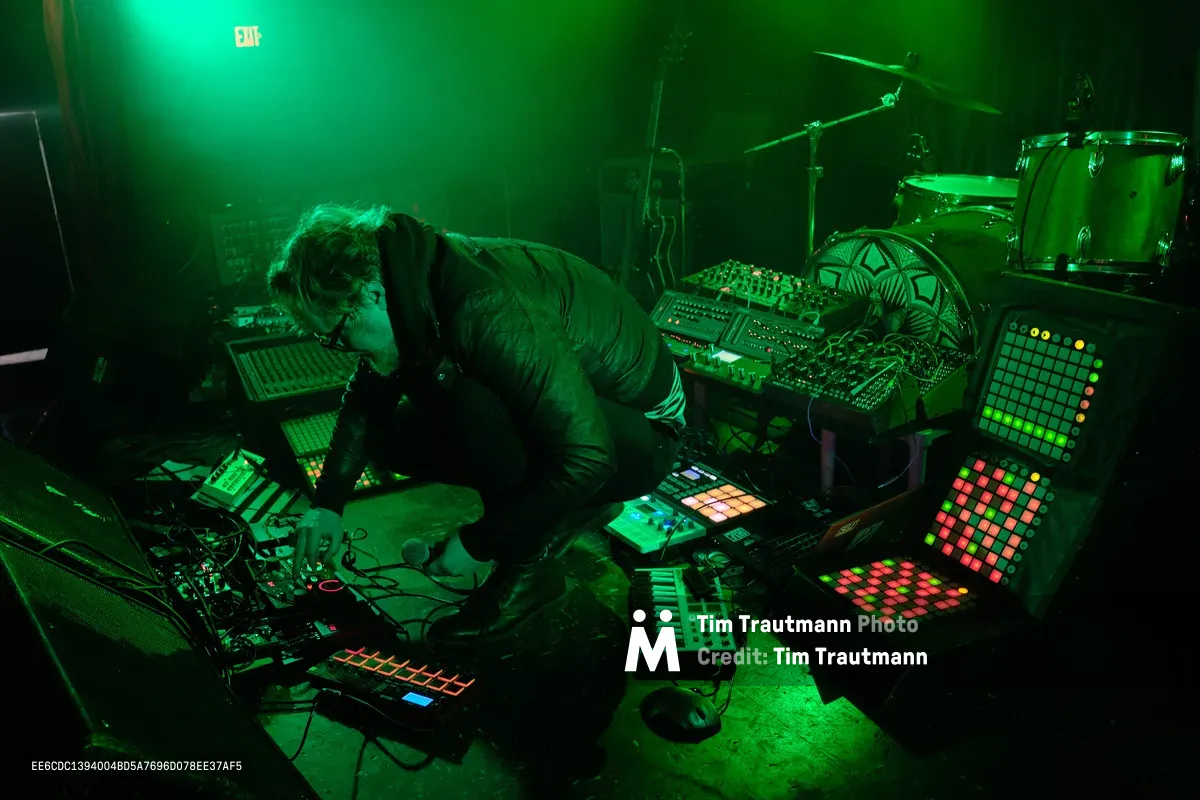 Bathed in supernatural emerald light, electronic musician Eli Savage hunches over his intricate array of synthesizers, drum machines, and glowing grid controllers during his ellipsism performance at RXFest 2019. The atmospheric green wash transforms Dante's intimate venue into a cybernetic cathedral, where analog knobs and digital displays pulse with multicolored frequencies. Savage's concentrated posture suggests deep communion with his electronic arsenal, while the moody lighting creates dramatic shadows that blur the line between performer and machine.