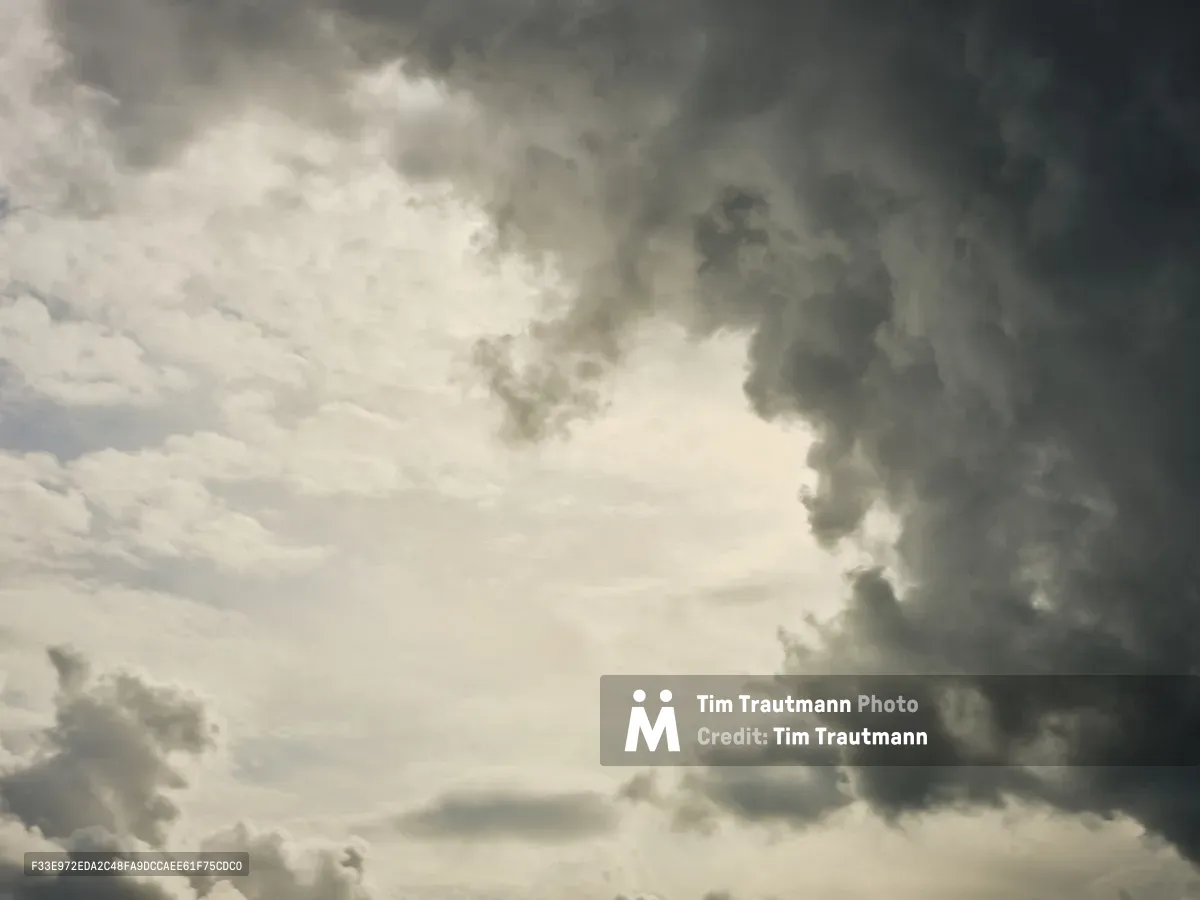 Brooding cumulonimbus clouds mass in pewter and charcoal tones above Portland's Central Eastside district, their heavy forms caught in dramatic chiaroscuro against patches of luminous sky. The monochromatic palette emphasizes the raw power of the approaching weather system, with billowing cloud formations creating a sense of impending atmospheric drama over the city's industrial corridor. Light breaks through in ethereal beams, casting the entire skyscape in theatrical contrast between shadow and illumination.