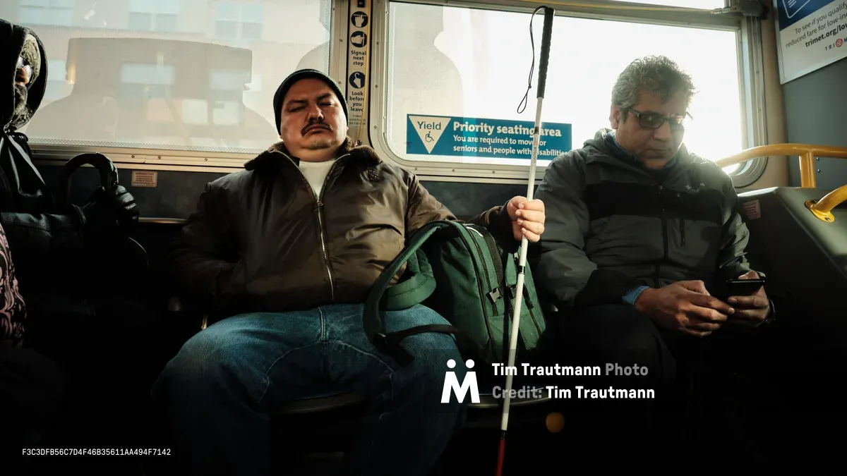 Golden morning light streams through the windows of a TriMet bus as it carries commuters through Northeast Portland's historic Eliot district. A passenger with a white cane sits peacefully in the priority seating area, while another commuter checks his phone, both bundled against the Pacific Northwest chill in dark winter jackets. The warm interior glow contrasts with the pale overcast sky visible through rain-spotted windows, capturing the quiet rhythm of urban transit accessibility.